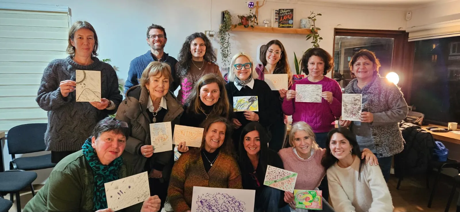 Un grupo de mujeres y un hombre posan sonrientes en una sala, en tres filas, cada persona mostrando el dibujo que hizo en el taller.