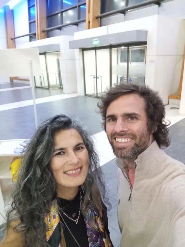 Maca e Ignacio sonríen a la cámara en una autofoto, dentro de un edificio con ventanales.