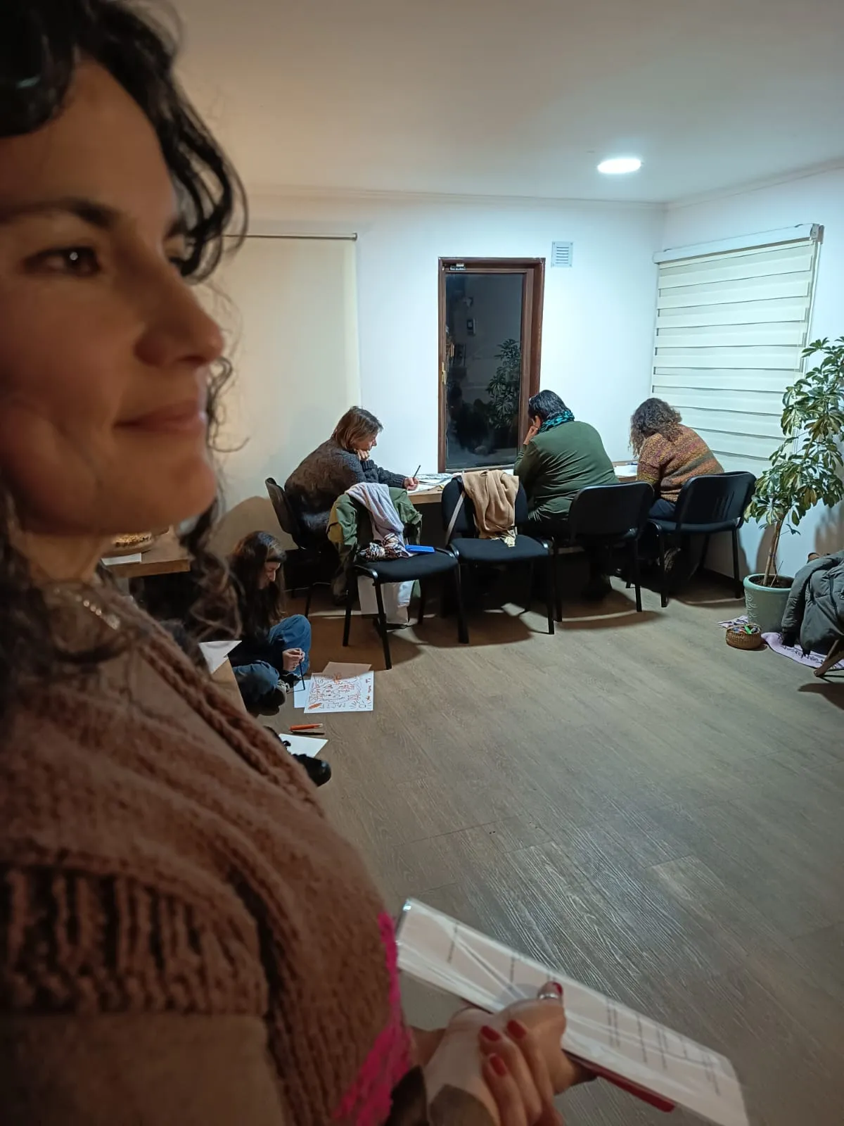 Maca, de perfil y sonriendo, con una libreta en la mano; al fondo, varias personas dibujan en una sala.